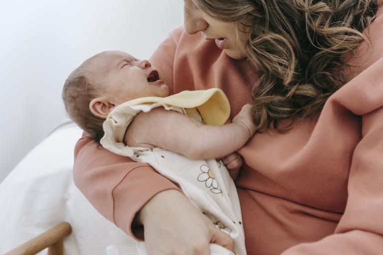 Side view of crop anonymous mother consoling crying infant baby in arms while sitting in light room near wall at home